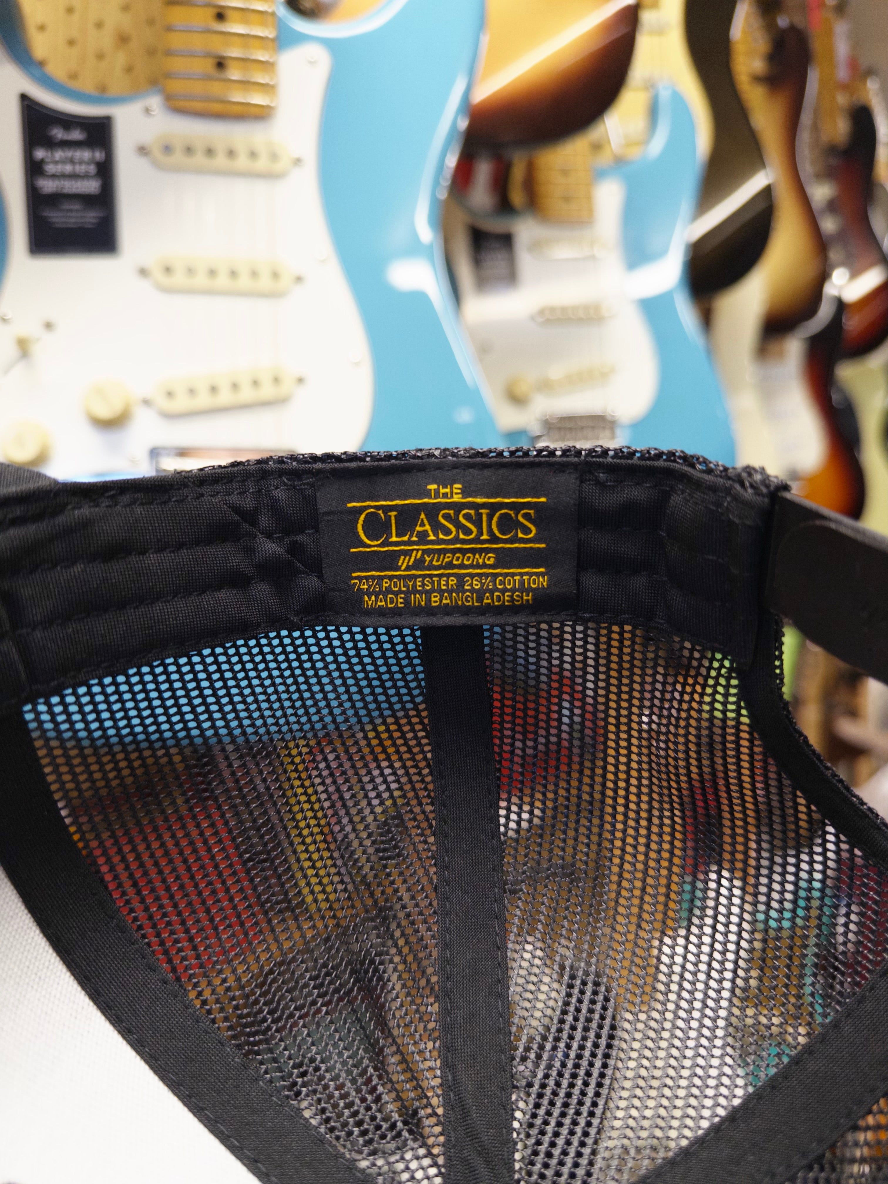 Close-up of a cap with 'The Classics' branding, blurred guitars in the background