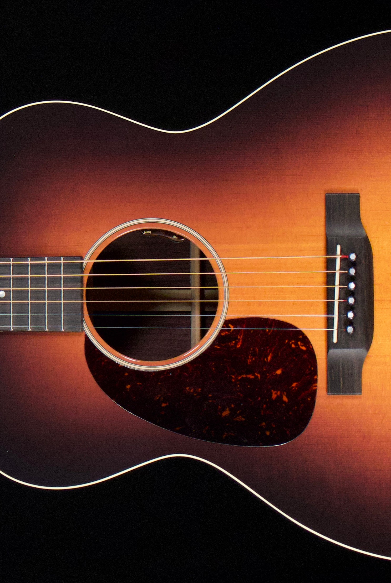 A close-up of an acoustic guitar body showing a classic sunburst finish that transitions from black at the edges to a warm amber-orange center, with a dark tortoiseshell pickguard and white binding.