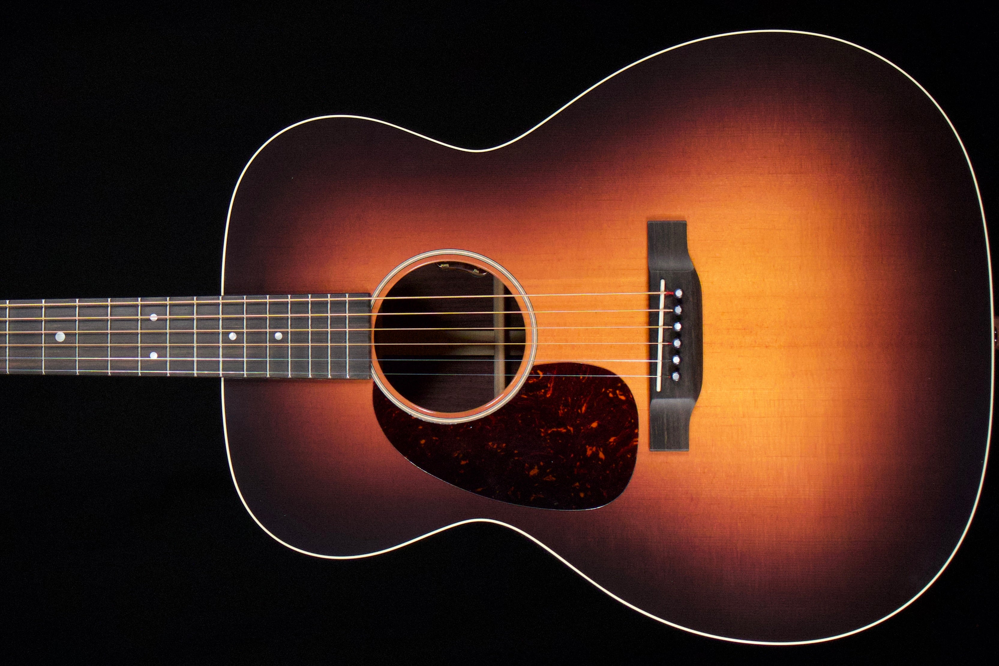 A close-up of an acoustic guitar body showing a classic sunburst finish that transitions from black at the edges to a warm amber-orange center, with a dark tortoiseshell pickguard and white binding.